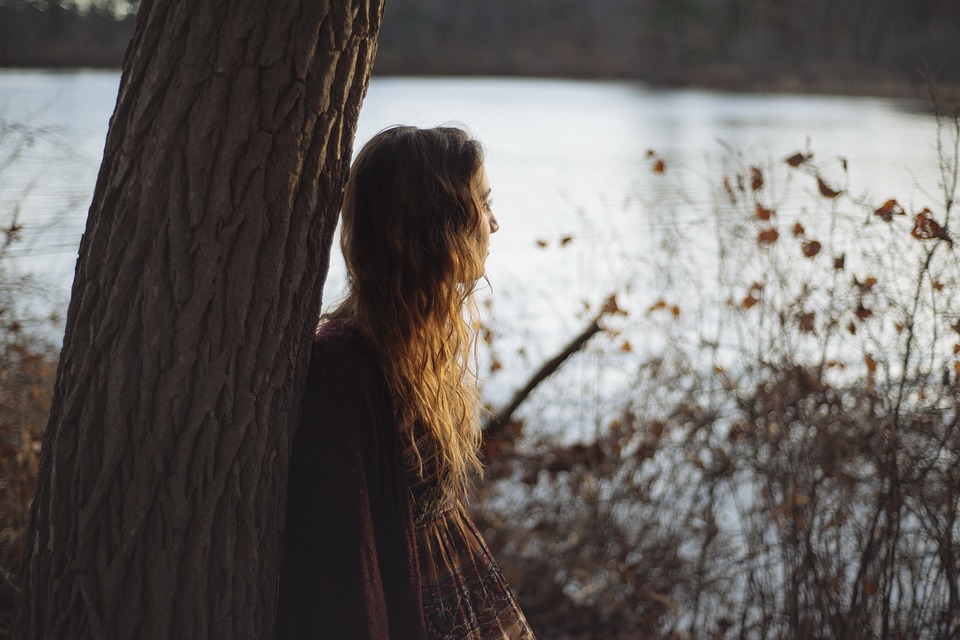 A young woman leaning against a tree staring out at a pond.