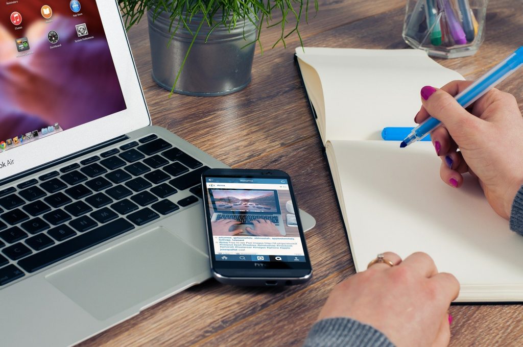 A laptop, mobile phone, and notepad sitting on a desk. Hands are writing in the notepad with a blue pen.