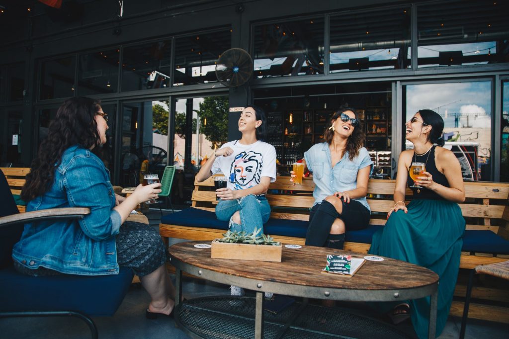 Four young women sitting outside a bar laughing and drinking beers.