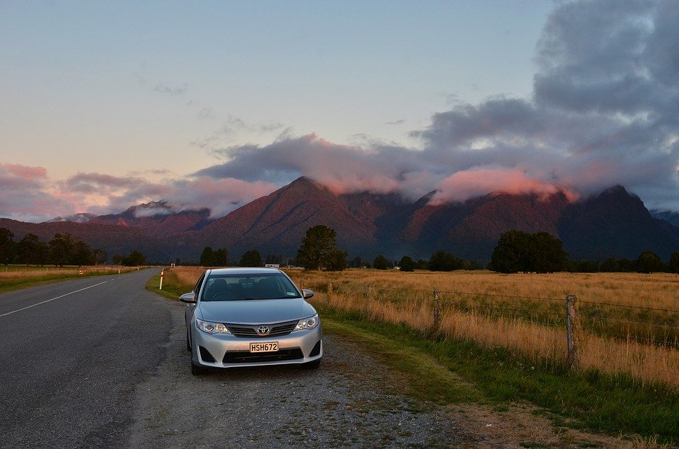 New Zealand mountains at sunset with a road and field in the foreground. A silver car is pulled off the road in a turnoff.