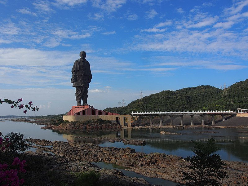 The Statue of Unity in the distance stands on top of a building with a bridge leading in from the right side. A blue sky and some hills lie behind the statue with a river and some rocks in front of it.