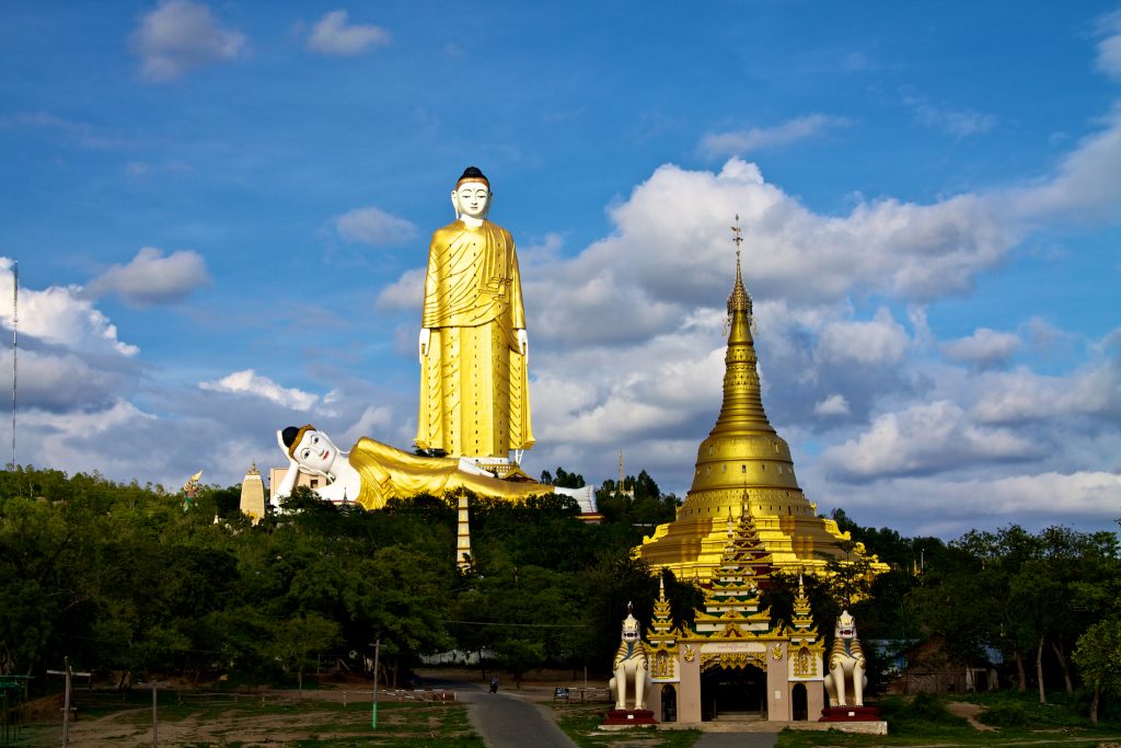 A distance photo of the yellow and white Laykyun Sekkya statue of Buddha and the second reclining Buddha at its feet. Trees and a golden-roofed temple in the foreground.