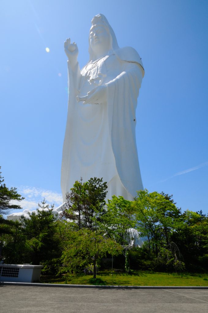 Looking up at the tall, white Sendai Daikannon statue. Blue sky behind and trees at its feet.