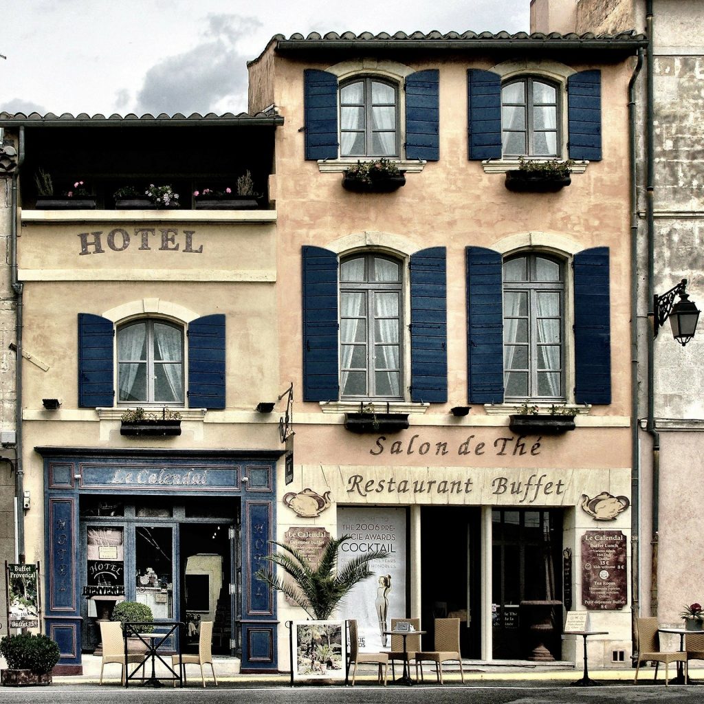 A quaint three-story hotel with arched windows and black shutters. A Salon de The and Restaurant Buffet are on the ground floor with doors open to a sidewalk with several small tables.