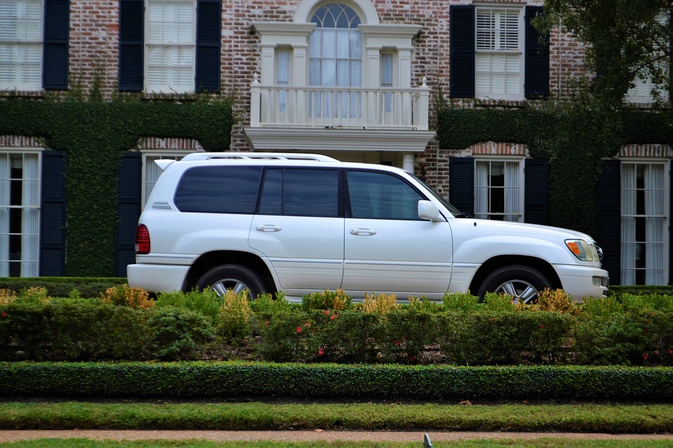 A white SUV parked in front of a larger brick house. Some shrubbery in the foreground.