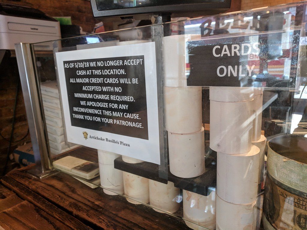 Clear plastic screen at a cafe counter with signs reading "Cards Only" and "As of 5/10/18 we no longer accept cash at this location. All major credit cards will be accepted with no minimum charge required. We apologize for any inconvenience this may cause. Thank you for your patronage."