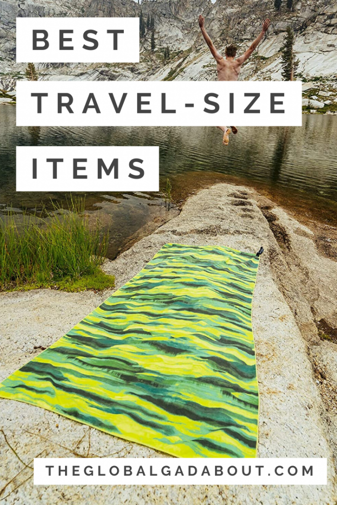 A green microfiber travel towel on the rocky shore of a lake with a man jumping into the lake in the distance. White rectangles overlaying the image with black words reading, "Best Travel-Size Items" and "TheGlobalGadabout.com".