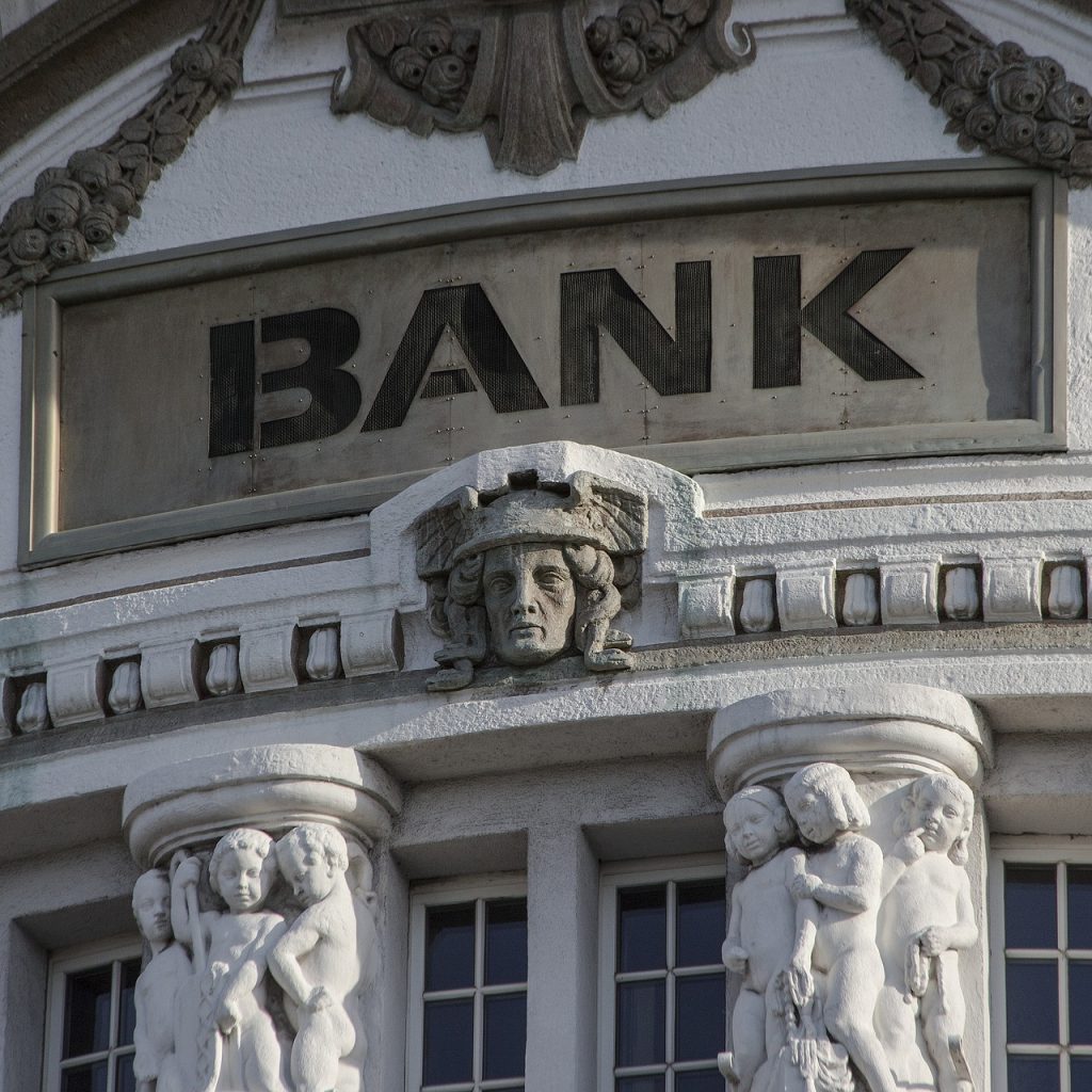 Close up of part of a building with the tops of two pillars, several carved figures & faces and the word "Bank".