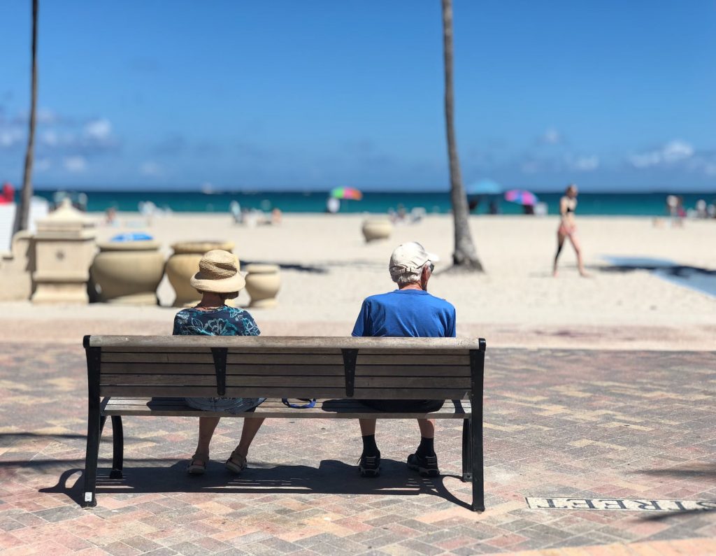 Beachside boardwalk with a bench. An older couple are seated on the bench looking at the water with their backs to the camera.