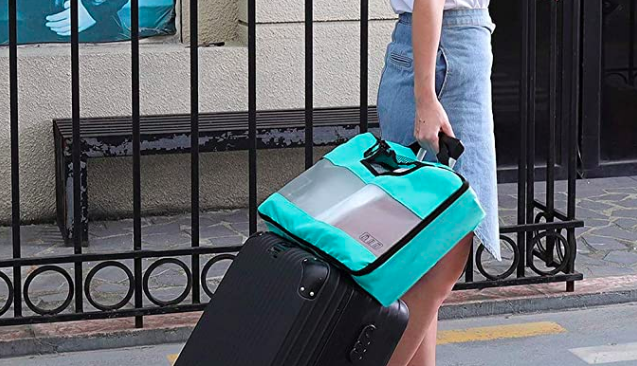 A woman walking down a street with a rolling suitcase and a packing cube stacked top, leaning against the suitcase handle.