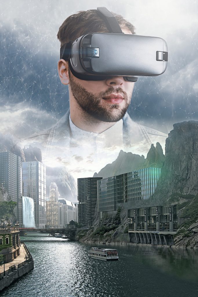 A river with a ferry and skyscrapers set into the cliffs on both sides of the river, a bridge and waterfall in the distance. A man's face wearing a virtual reality headset is floating in the sky, which has looks slightly digital.