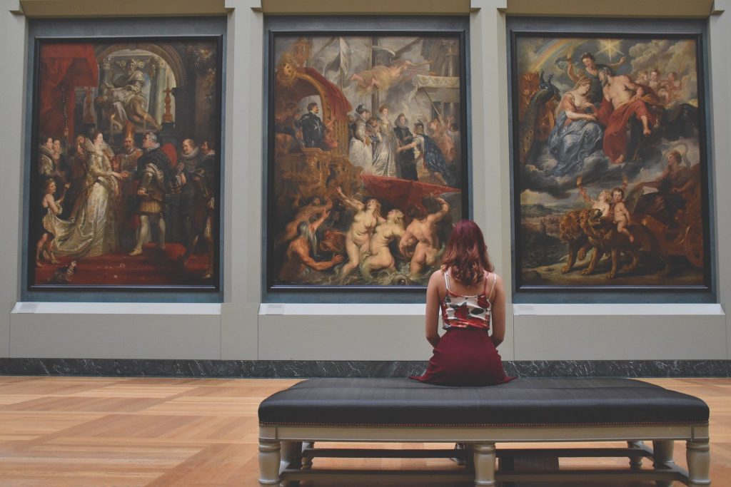 Three large paintings in the Louvre Museum with a woman sitting with her back to camera looking at the paintings.