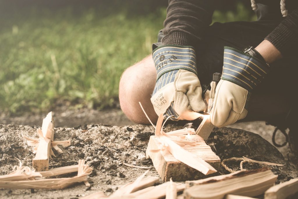 A pair of hands in work gloves spark flint to light a fire. Wood and ashes are in the fire pit.