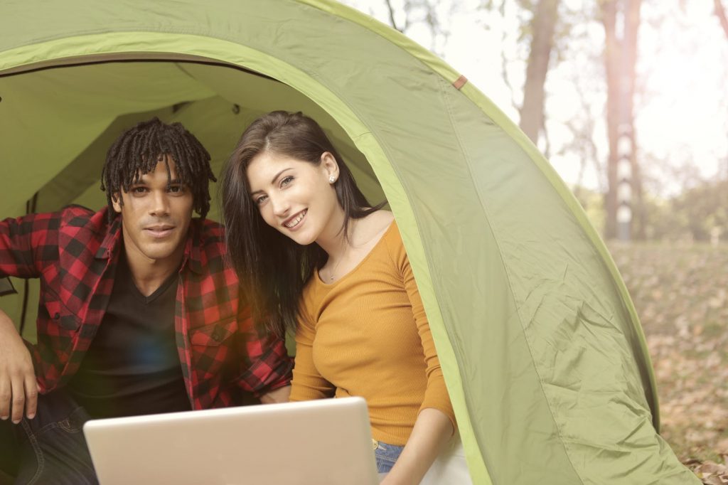 A close up of the opening of a tent. A young man and woman look out the open tent door with a laptop in front of them.