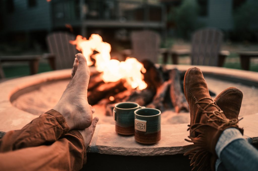 A campfire in a fire pit with chairs in the background. Two sets of crossed feet and two mugs are in the foreground.