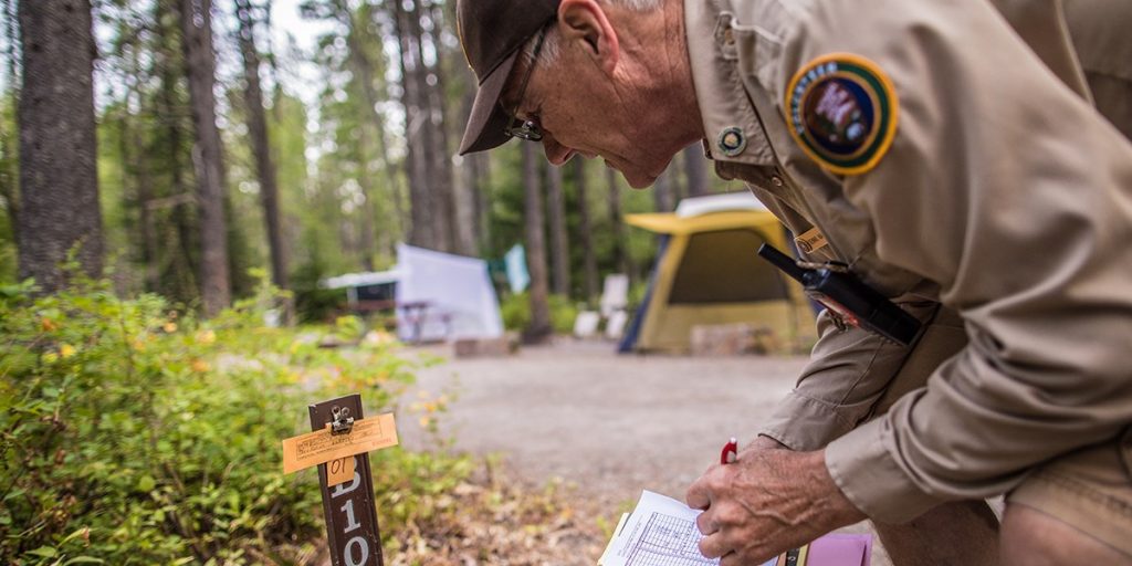 An older man in uniform leans over at a campsite and marks something on a paper in his hand.