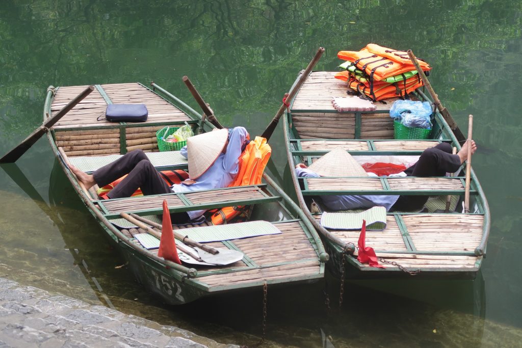 2 Asian row boats floating in shallow water. A person is asleep in each one with a conical hat covering their faces.