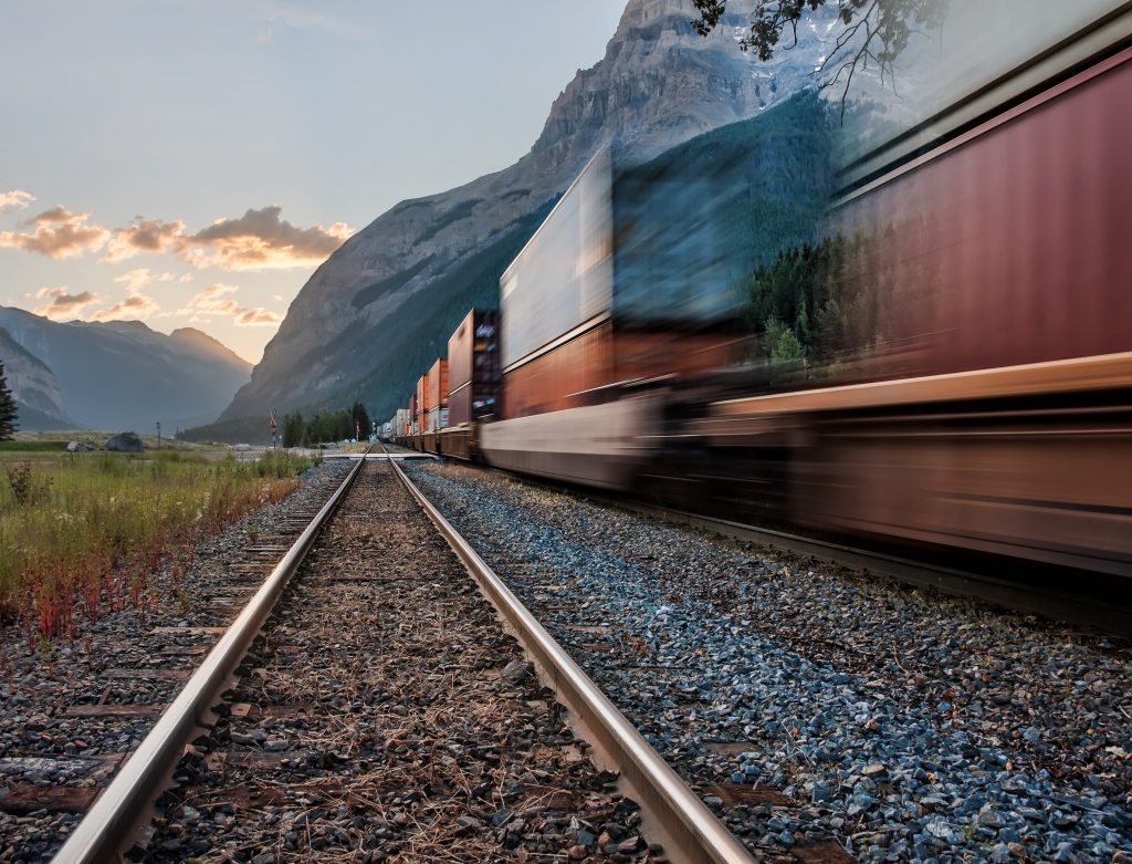 From the low perspective of the tracks, a train blurred by motion is heading toward the sun setting behind distant mountains.